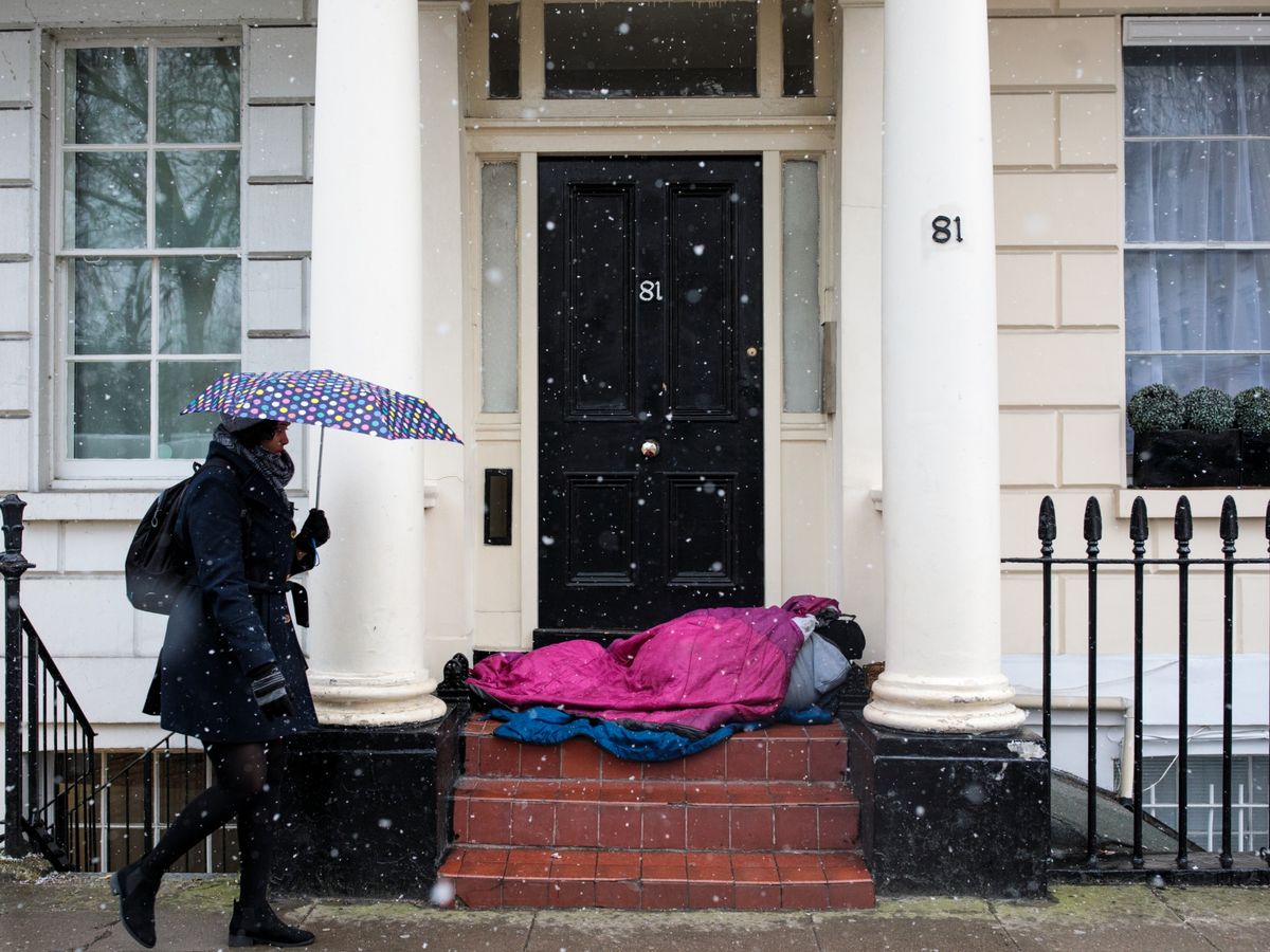 A-woman-carrying-an-umbrella-walks-past-a-homeless-person-sleeping-in-a-doorway-during-a-snow-shower-on-February-26